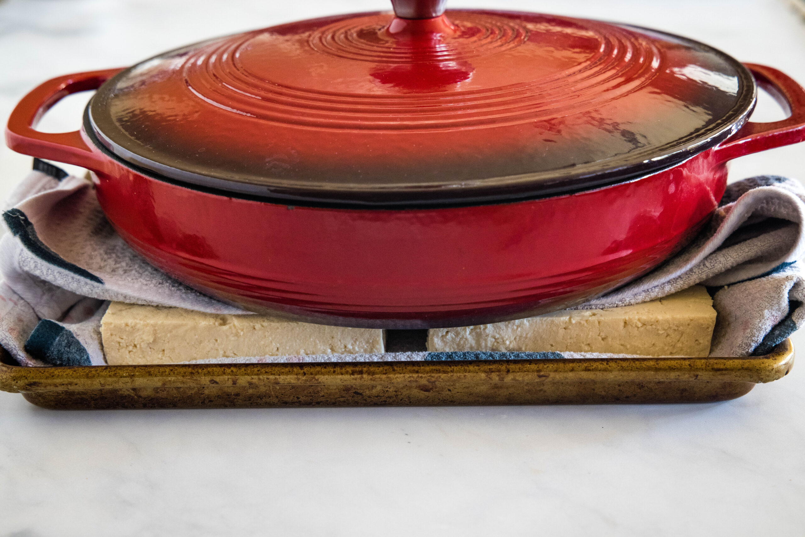 A large red skillet resting on top of tofu wrapped in kitchen towels, to press out the excess liquid.
