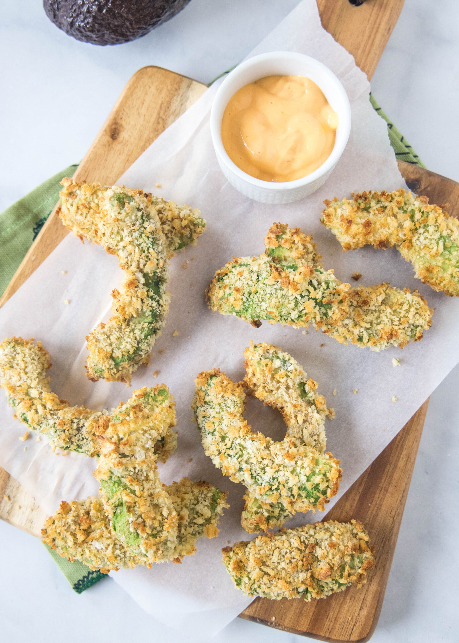 Overhead view of scattered avocado fries on a wooden cutting board lined with parchment paper, next to a small bowl of dipping sauce.