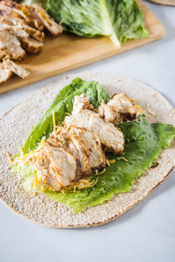 A tortilla wrap topped with a lettuce leaf, shredded cheese, and shredded chicken.