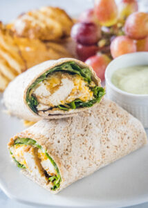 Close up of two halves of a Chick Fil A Cool Wrap on a plate next to a bowl of dipping sauce, grapes, and potato chips.