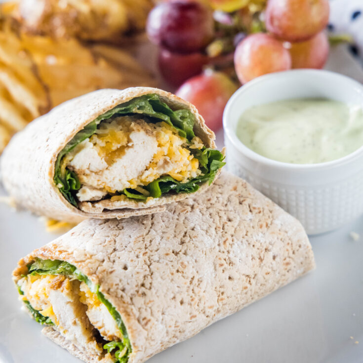 Two halves of a Chick Fil A Cool Wrap on a plate next to a bowl of dipping sauce, grapes, and potato chips.