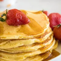 A stack of ricotta pancakes topped with a strawberry and maple syrup on a plate, next to more fresh strawberries.