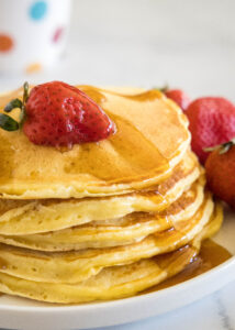 A stack of ricotta pancakes topped with a strawberry and maple syrup on a plate, next to more fresh strawberries.