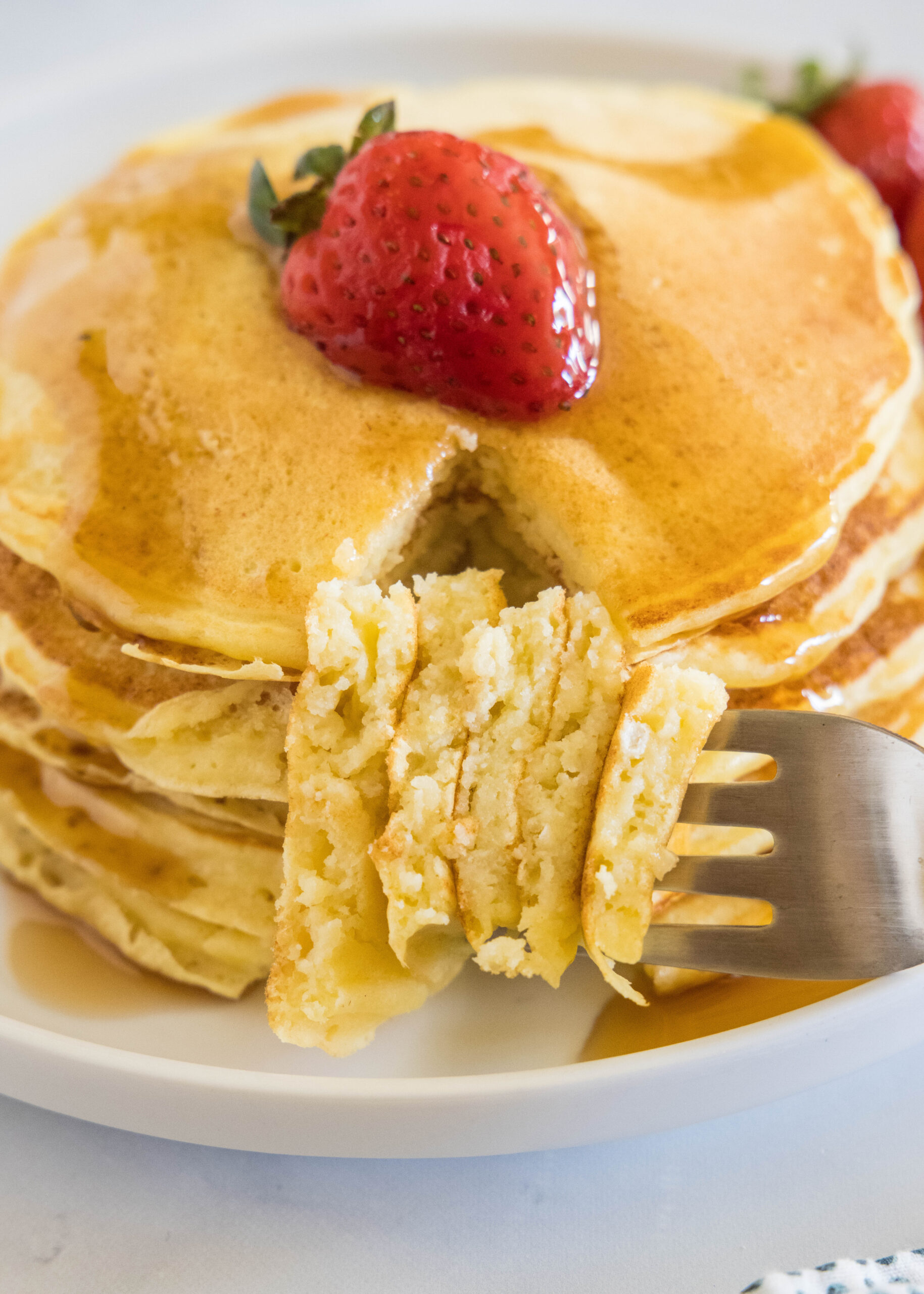 A forkful laying next stack of ricotta pancakes topped with a strawberry and maple syrup on a plate.