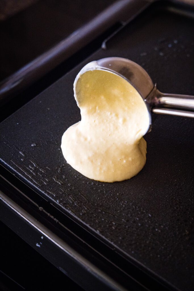 An ice cream scoop dropping a scoop of ricotta pancake batter onto a hot griddle.