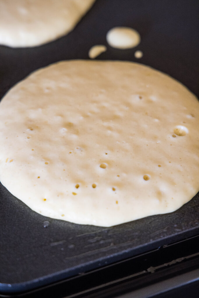 Ricotta pancake batter cooking on a griddle, showing small bubbles on the surface to indicate that it's ready to flip.