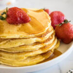 A stack of ricotta pancakes topped with a strawberry and maple syrup on a plate, next to more fresh strawberries.
