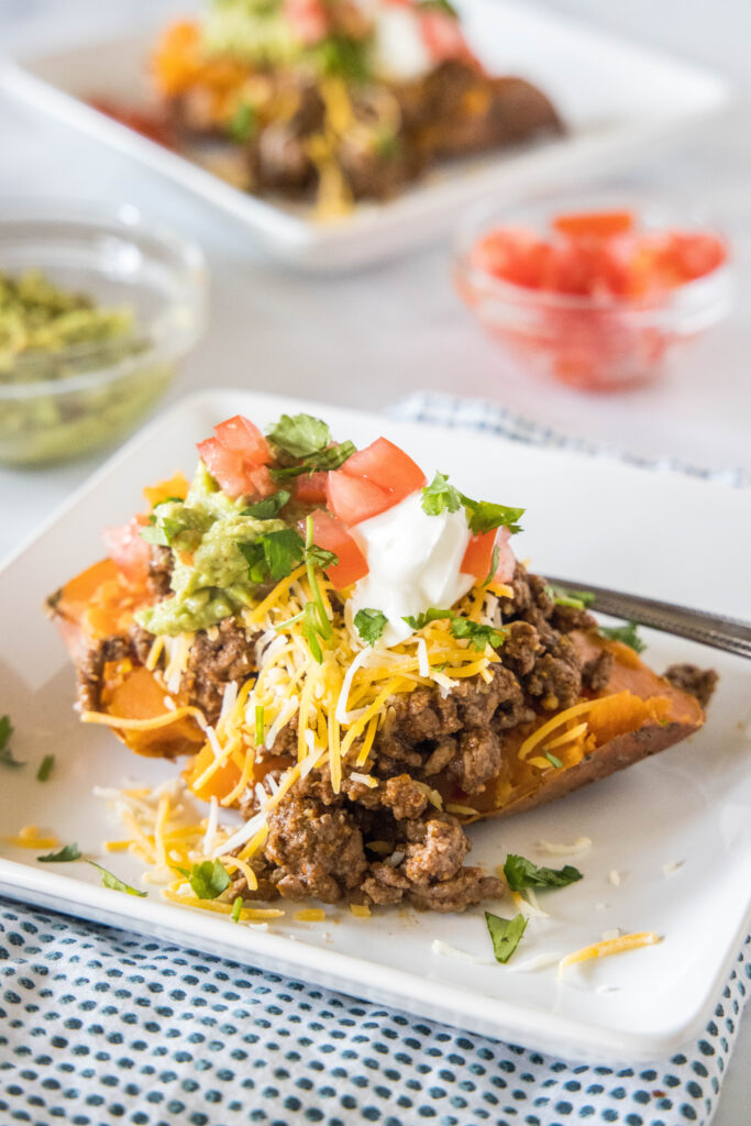 A taco sweet potato piled with taco toppings served on a white plate, with a second sweet potato and bowls of toppings in the background.