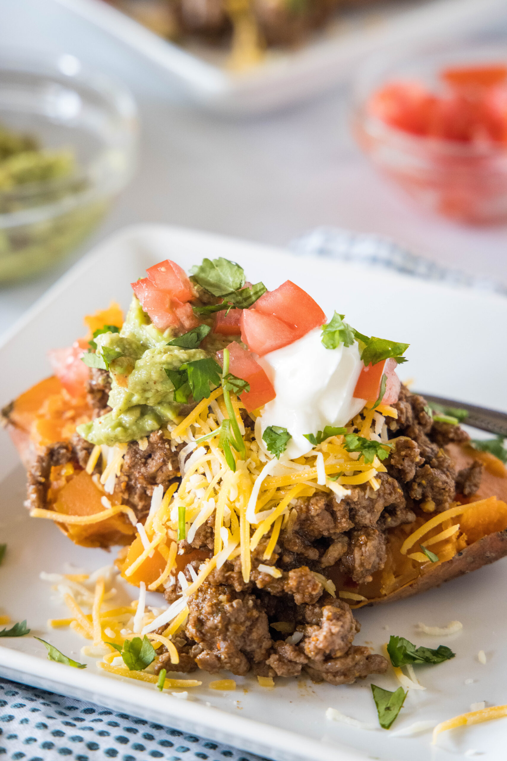 A taco sweet potato piled with taco toppings served on a white plate, with bowls of toppings in the background.