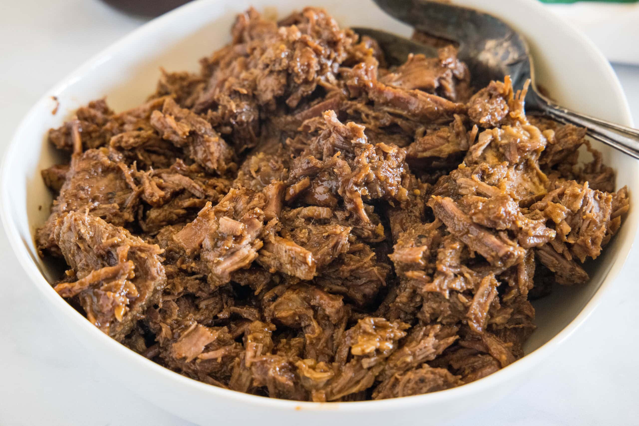 This BBQ beef is slow-cooked until it’s fall-apart tender and tossed with sweet-tangy barbecue sauce. Perfect for a potluck or cookout! Metal tongs resting in shredded BBQ beef inside the crock pot.