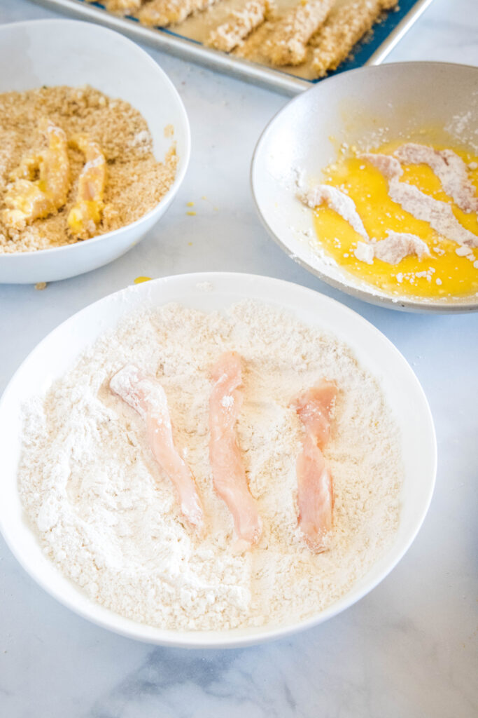 A breading station with chicken strips placed in bowls of flour, breadcrumbs, and egg wash.