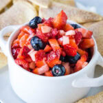 Fruit salsa in a white ceramic serving bowl with two handles, surrounded by cinnamon chips.