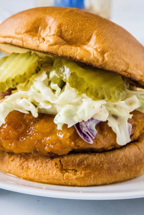 A hot honey chicken sandwich topped with coleslaw and pickles on a white plate.
