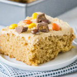 A slice of peanut butter cake topped with peanut butter frosting and chopped Reese's peanut butter cups on a plate.