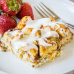 A serving of cinnamon roll casserole topped with glaze, next to a fork and fresh strawberries on a white plate.