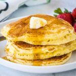 A stack of cornmeal pancakes topped with butter and maple syrup on a white plate.