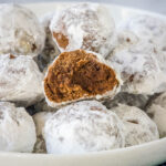 Chocolate snowball cookies staked on a white platter with a bite missing from one cookie.