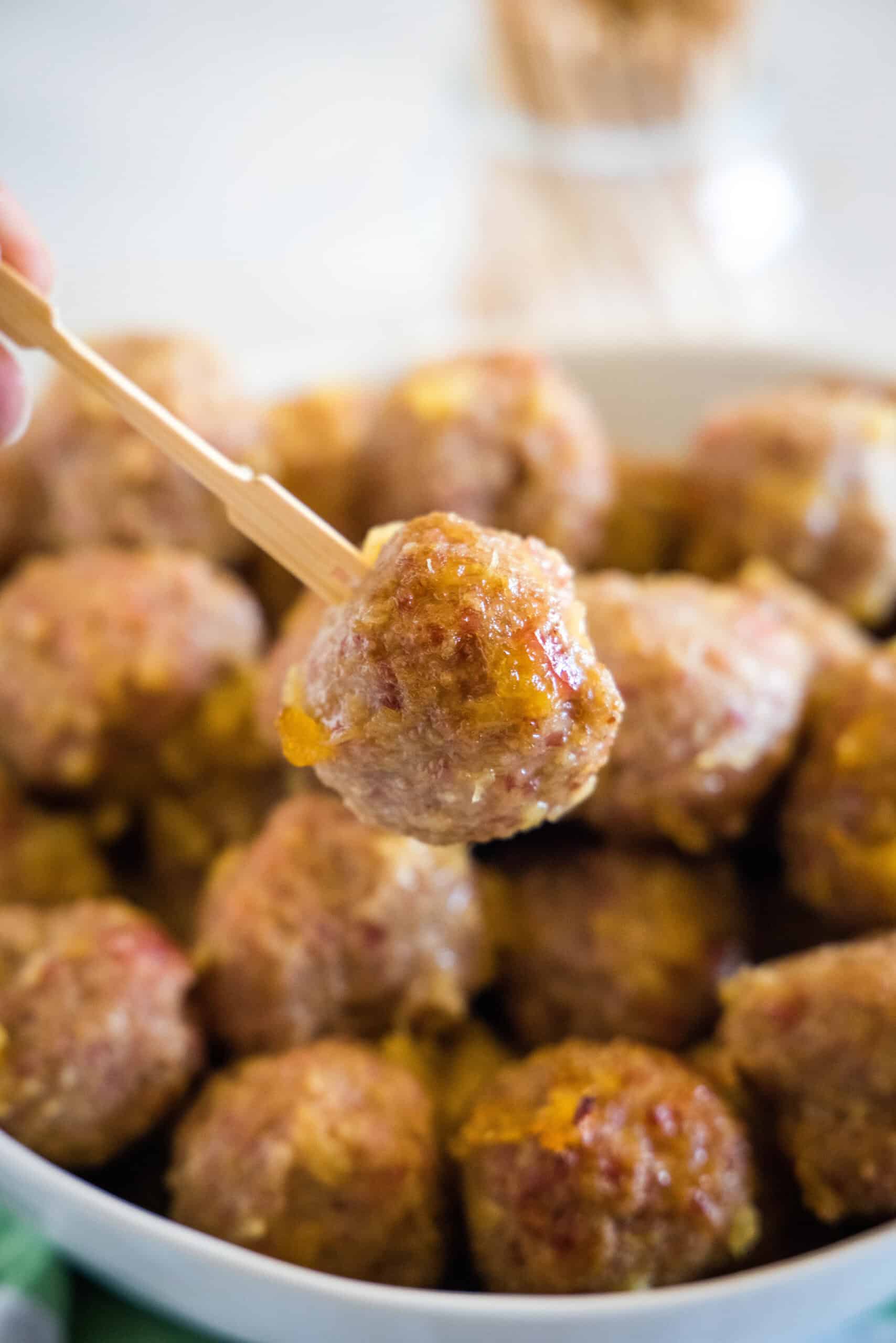 Close up of a ham ball picked up with a cocktail skewer, with a bowl of ham balls in the background.