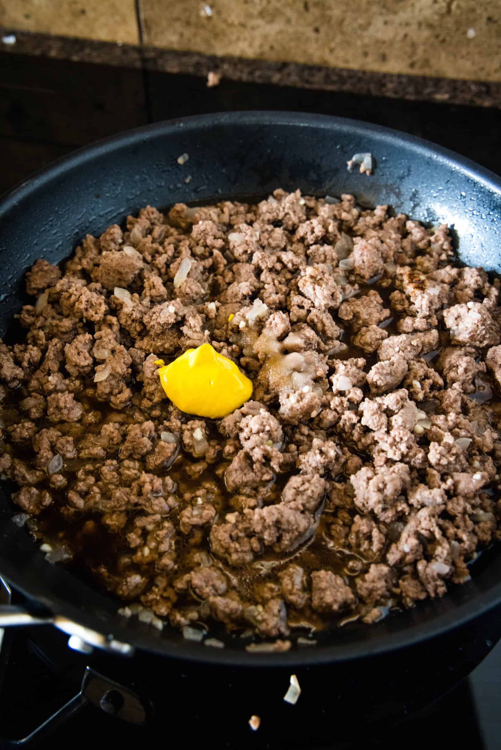 Mustard and seasonings added to browned ground beef in a skillet.