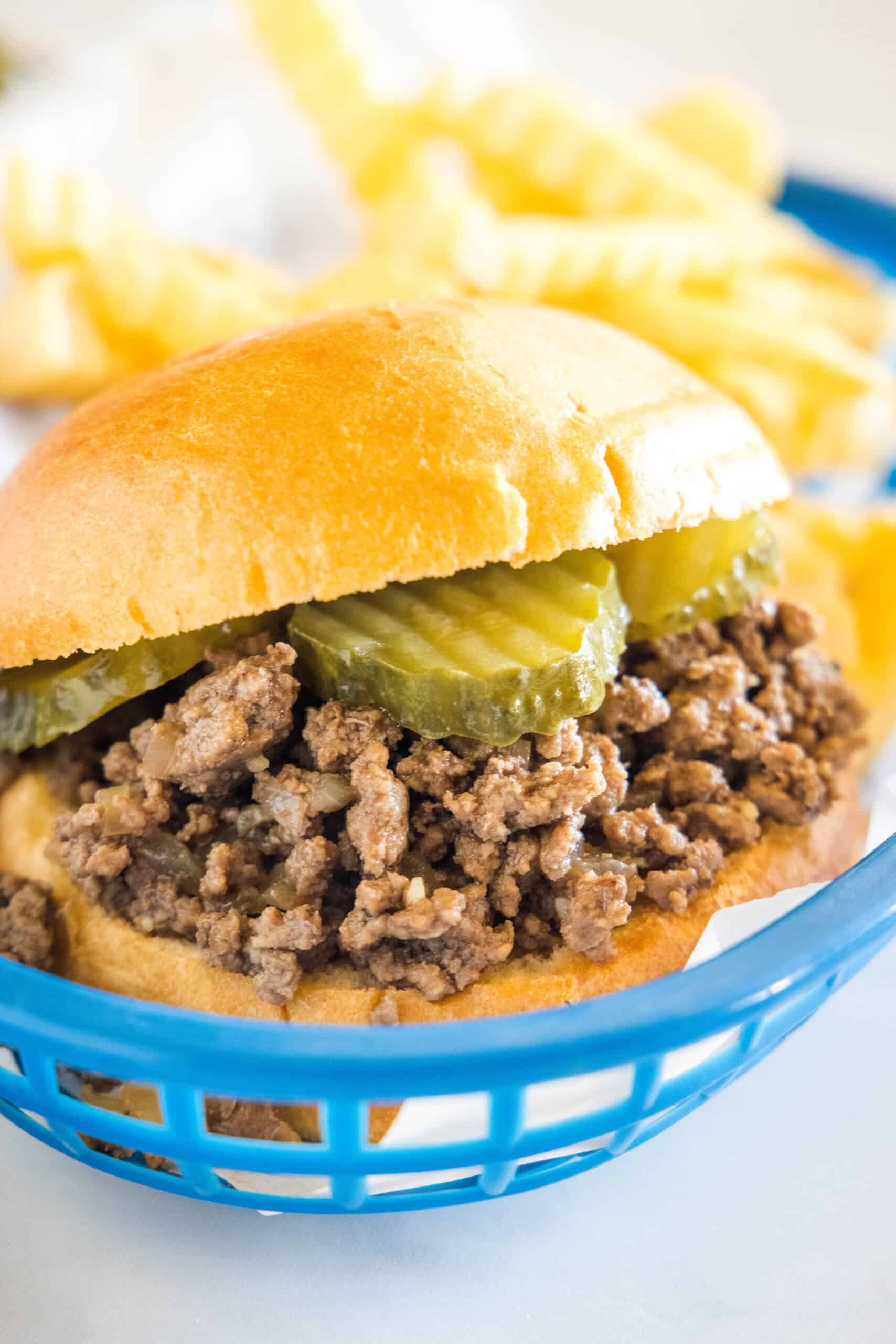 A Maid Rite sandwich garnished with pickles in a blue plastic serving basket, with fries in the background.