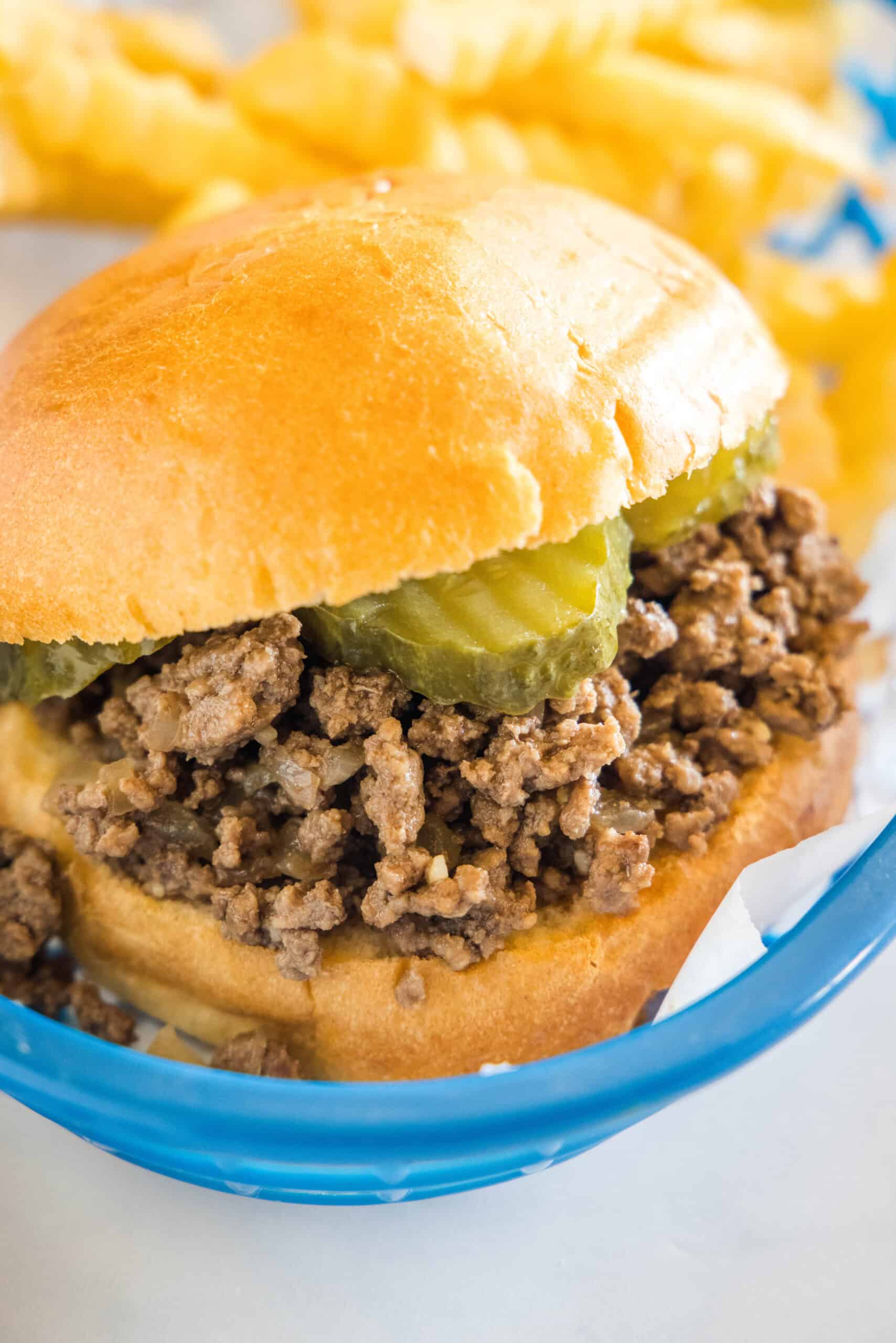 A Maid Rite sandwich garnished with pickles in a blue plastic serving basket, next to a side of fries.