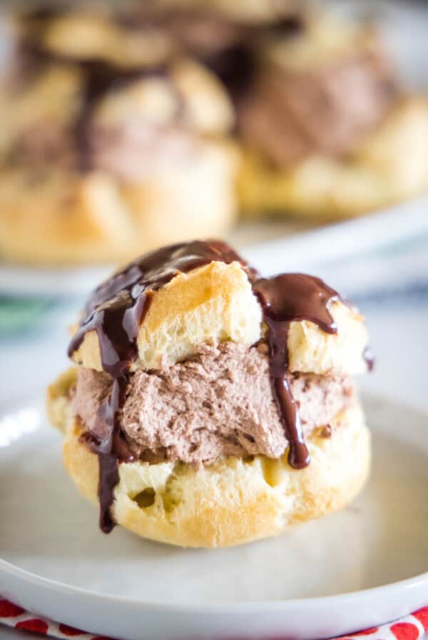 A chocolate cream puff topped with chocolate ganache on a white plate, with more cream puffs on a plate in the background.