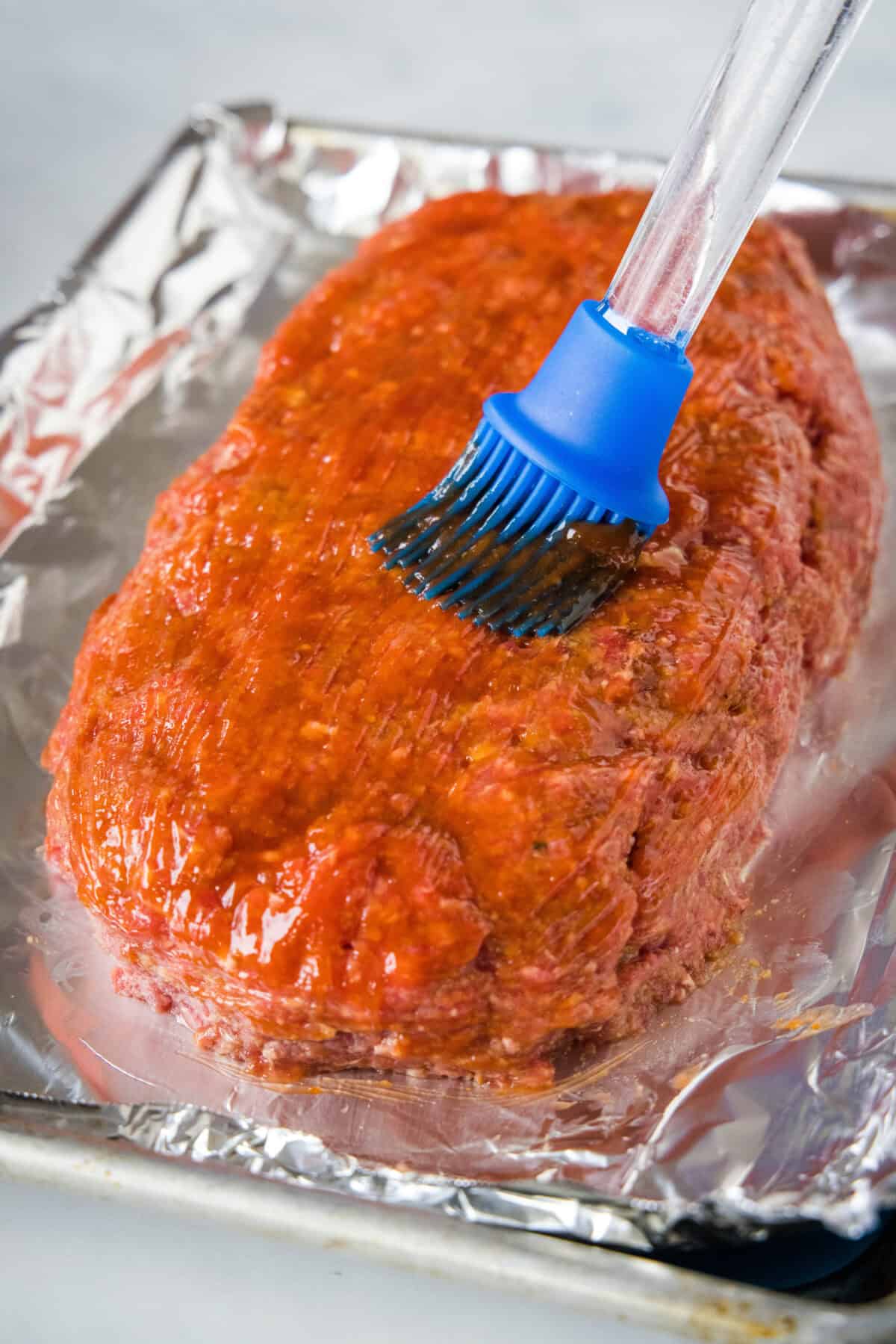 A blue basting brush brushing sauce over the outside of onion soup meatloaf on a lined baking sheet.