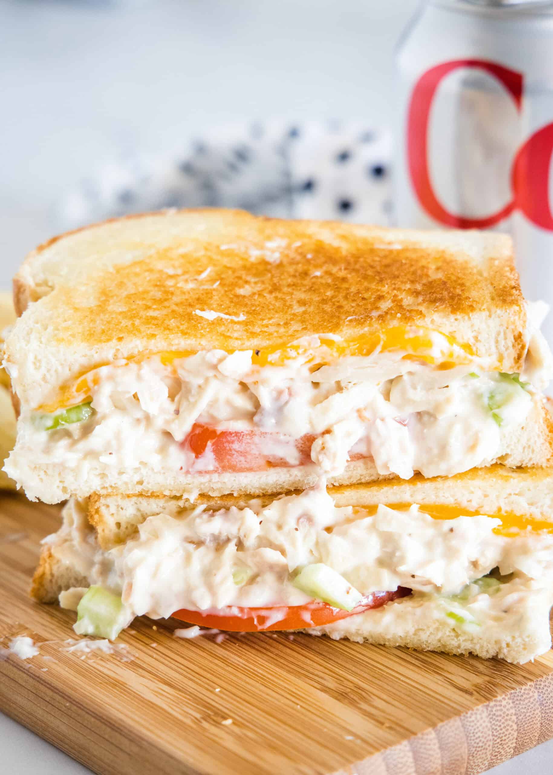 This is the best-ever tuna melt recipe with plenty of crunch, juicy tomatoes, and melty cheese between two golden, crispy bread slices! Two halves of a tuna melt stacked on top of one another on a wooden platter.