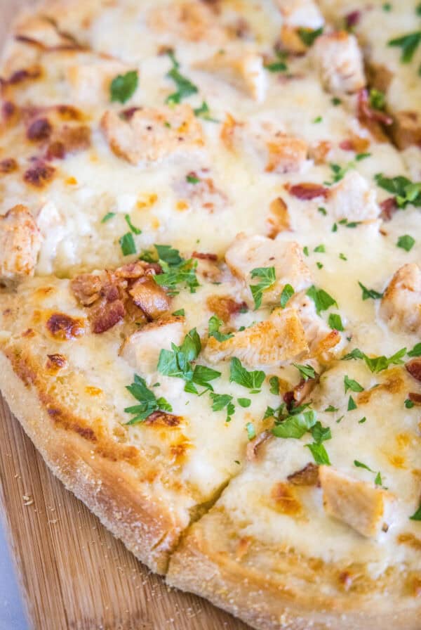 Close up of a chicken alfredo pizza cut into slices on a wooden butting board.