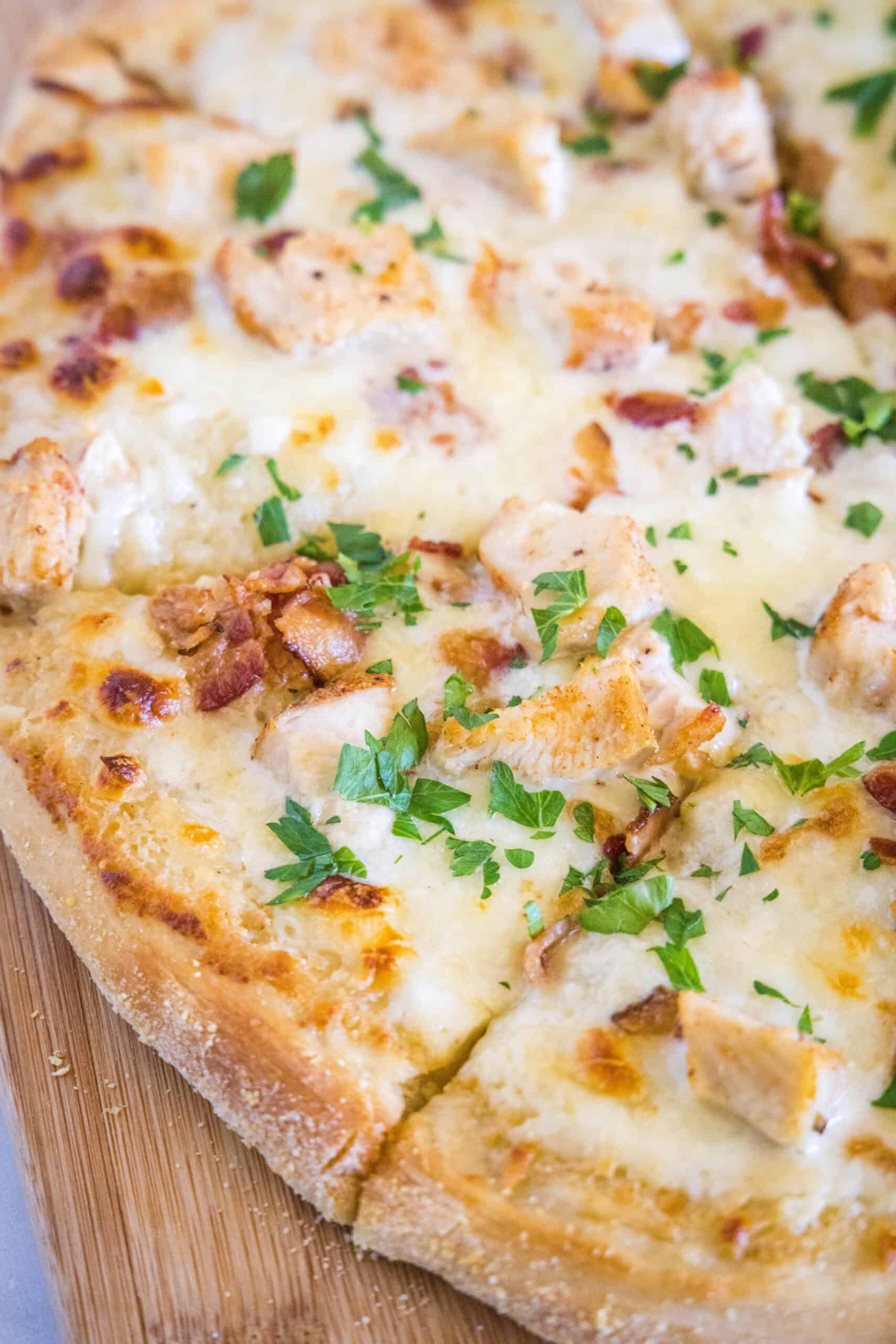 This chicken alfredo pizza has a white pizza base of creamy homemade alfredo sauce topped with juicy chicken, bacon, and melty mozzarella! Close up of a chicken alfredo pizza cut into slices on a wooden butting board.