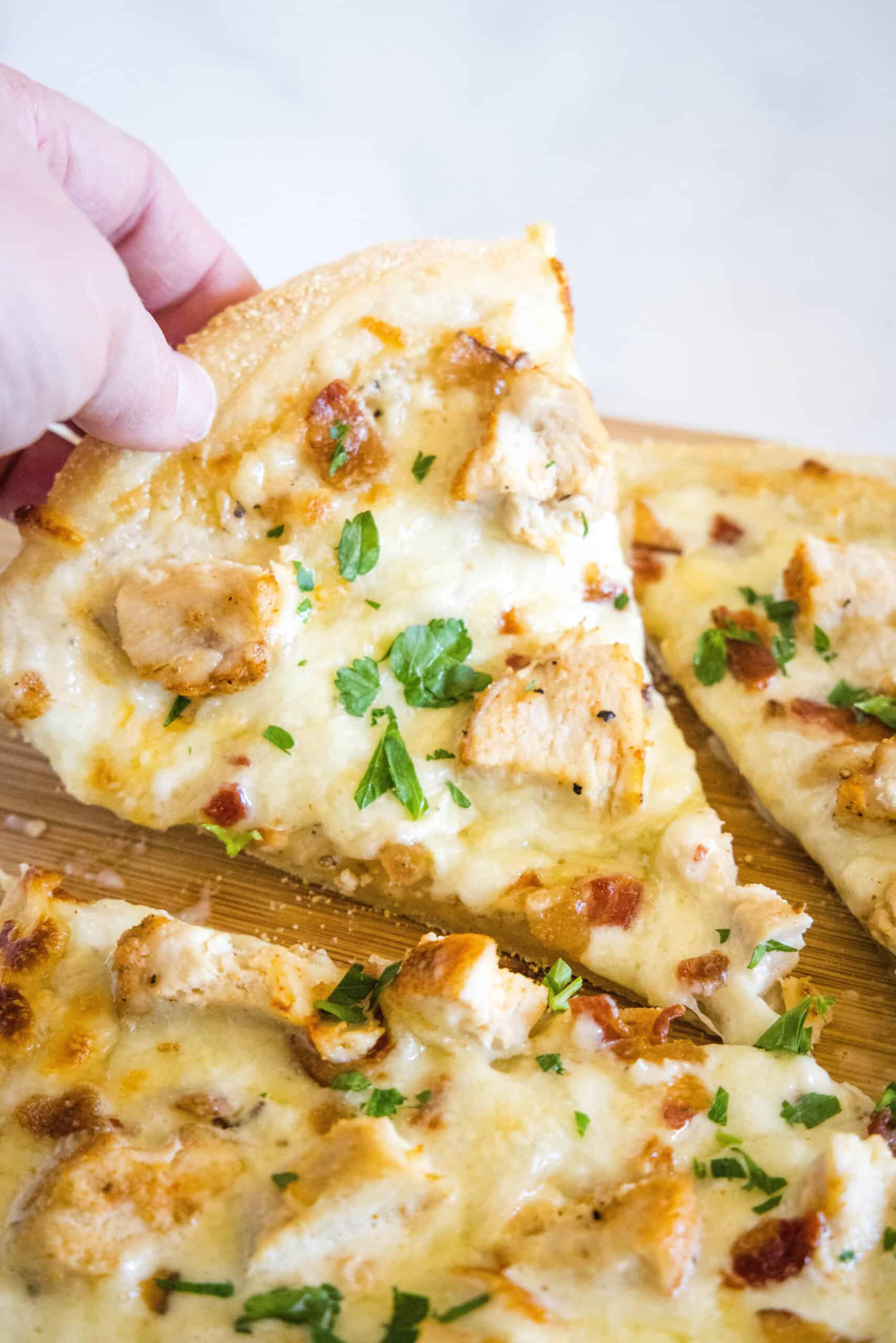 A hand lifting a slice of chicken alfredo pizza from a wooden cutting board.