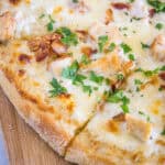 Close up of a chicken alfredo pizza cut into slices on a wooden butting board.