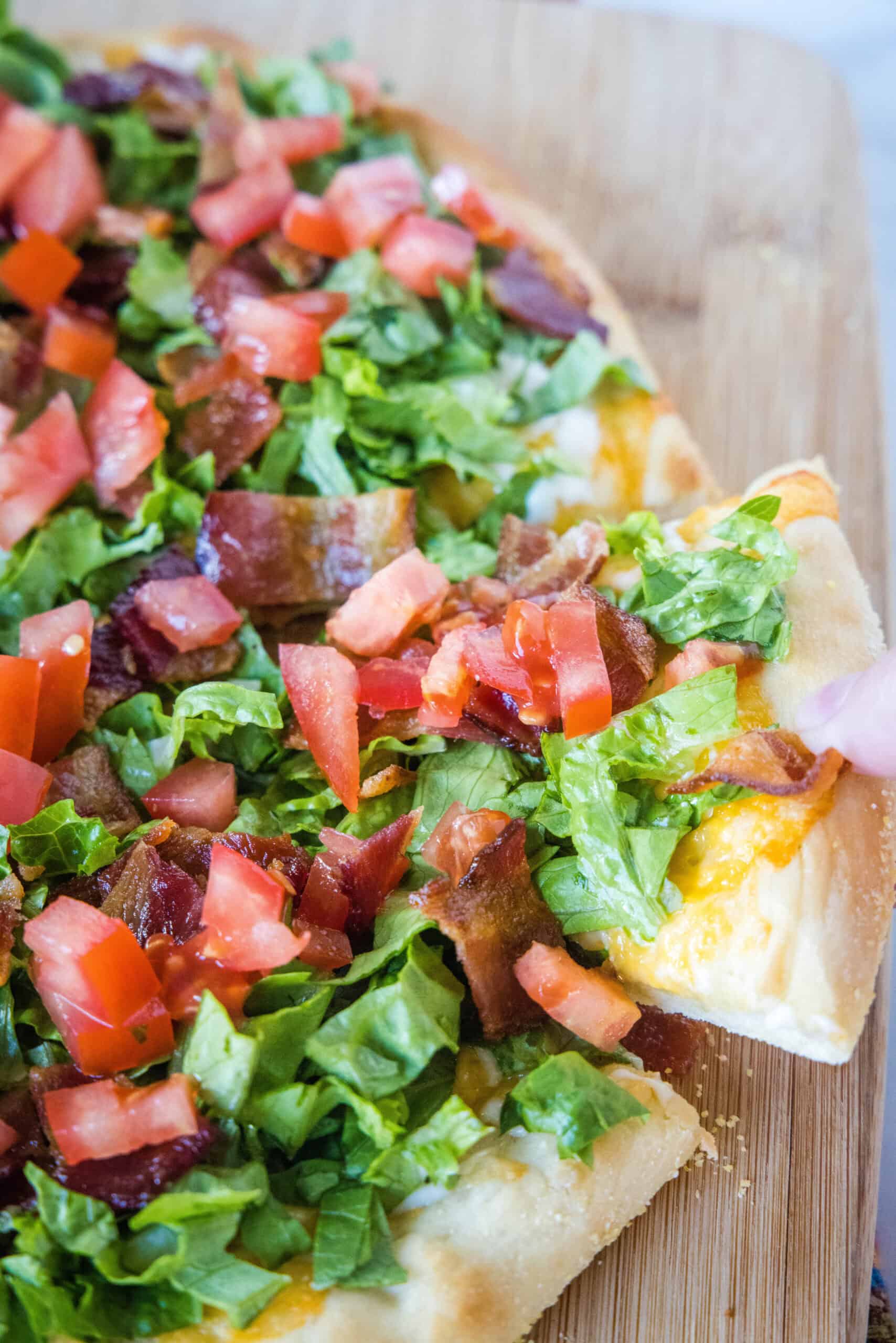 This BLT pizza recipe reimagines a classic bacon, lettuce, and tomato sandwich into a delicious, cheesy, and flavor-packed homemade pizza! A hand lifting a slice from a BLT pizza on a wooden cutting board.