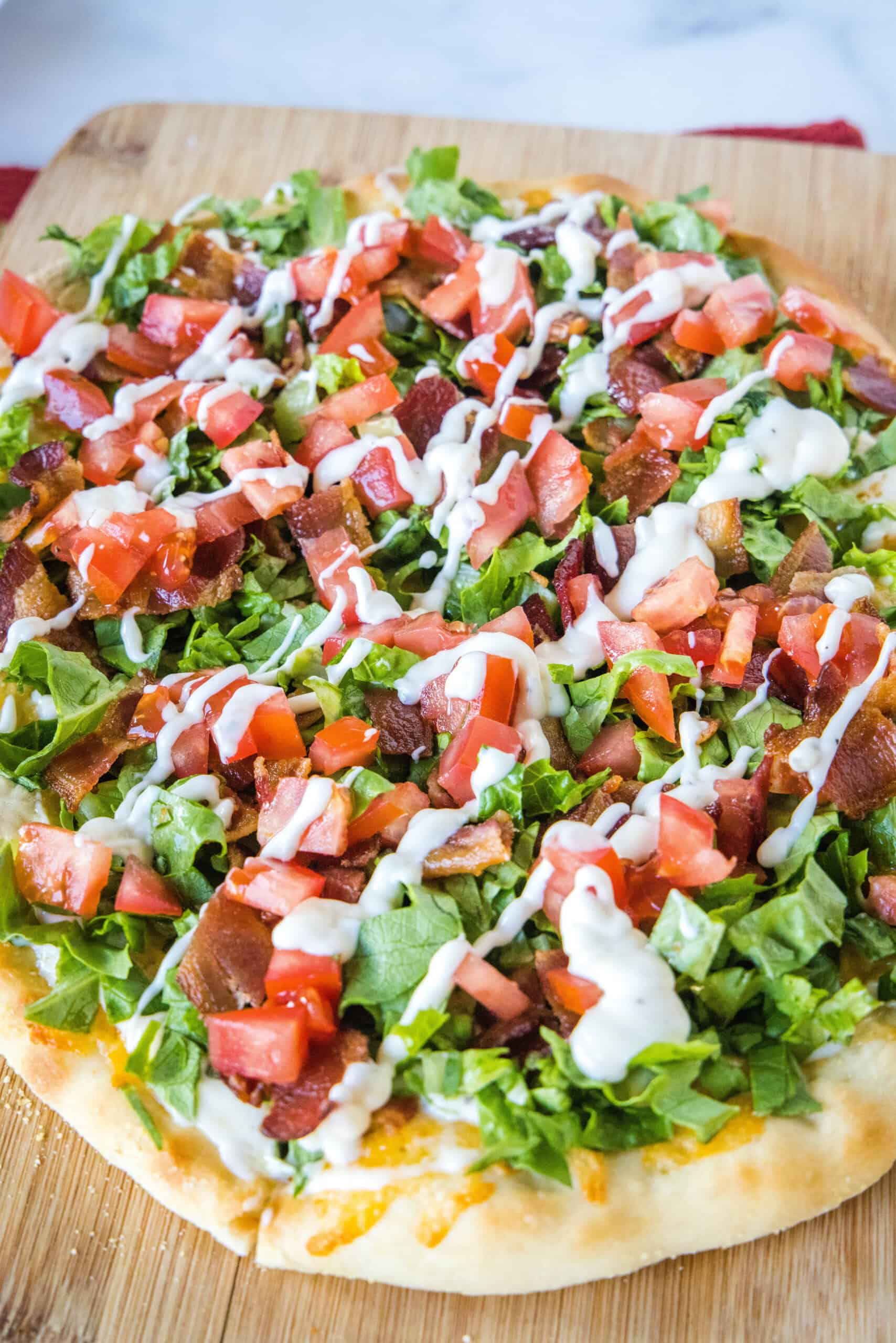 This BLT pizza recipe reimagines a classic bacon, lettuce, and tomato sandwich into a delicious, cheesy, and flavor-packed homemade pizza! Close up of a BLT pizza topped with a drizzle of aioli on a wooden cutting board.