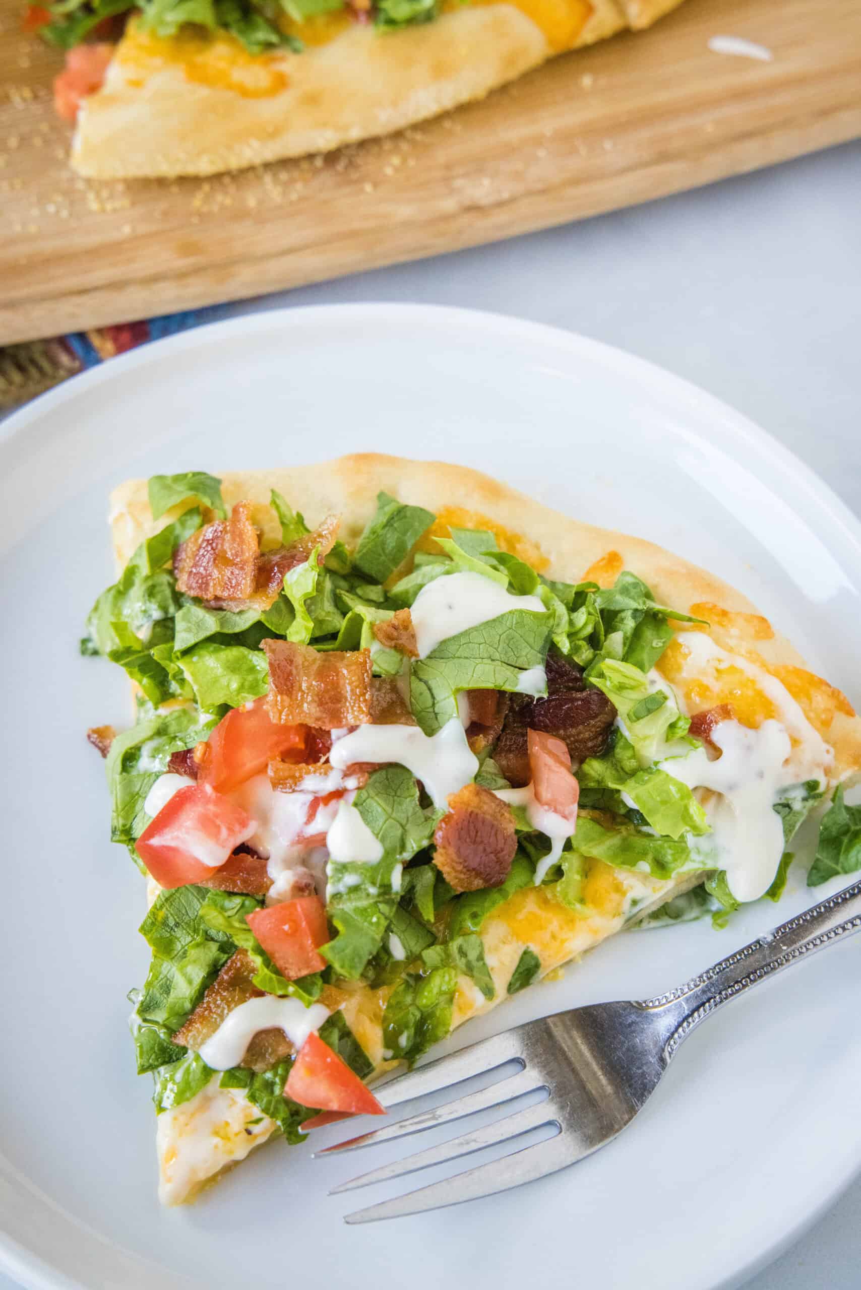 This BLT pizza recipe reimagines a classic bacon, lettuce, and tomato sandwich into a delicious, cheesy, and flavor-packed homemade pizza! A slice of BLT pizza on a white plate next to a fork.
