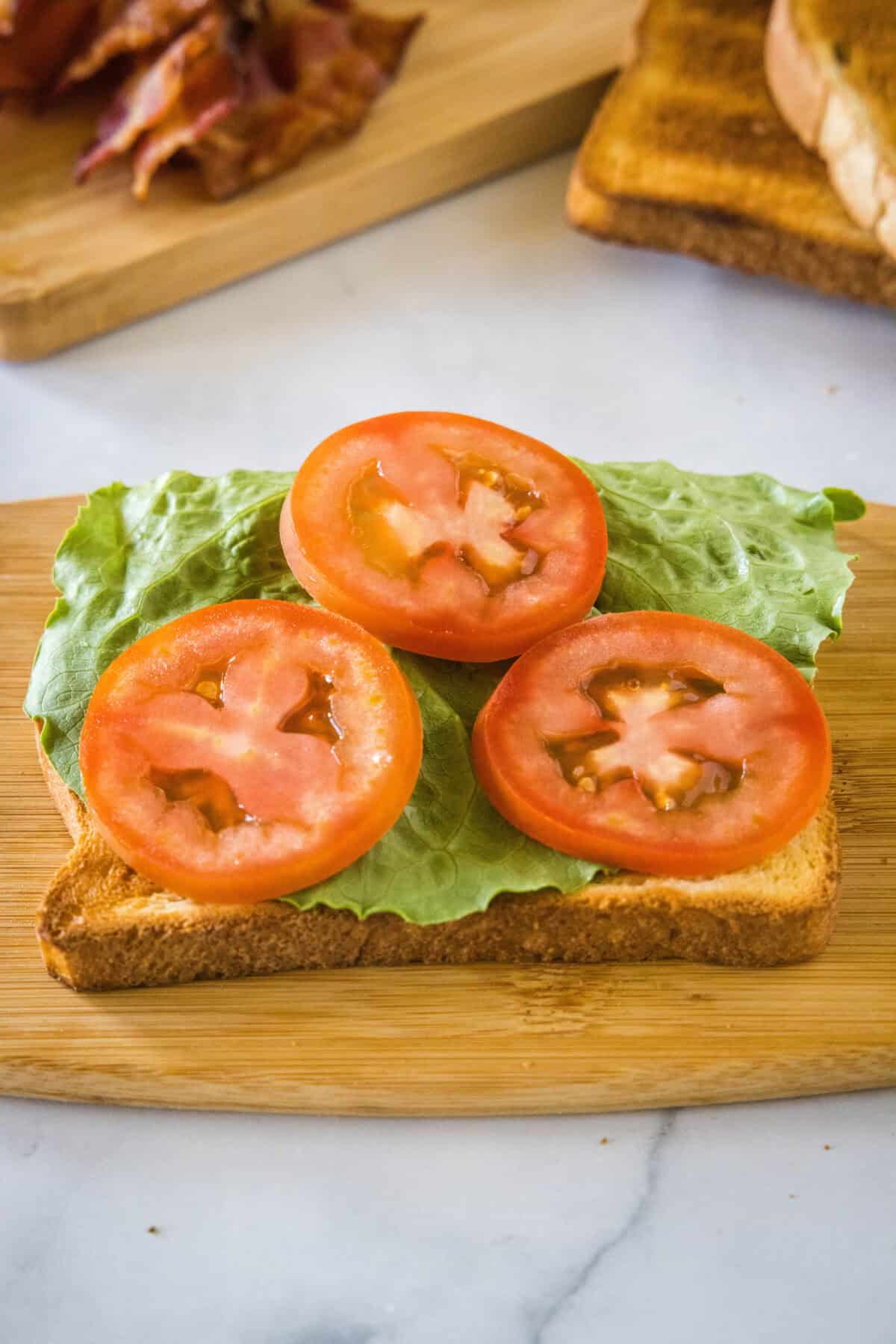 The first bread slice topped with lettuce and tomato on a wooden cutting board.