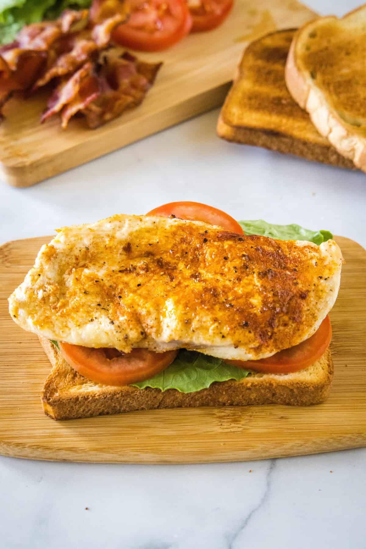 The first bread slice topped with lettuce, tomato, and a seared chicken piece on a wooden cutting board.