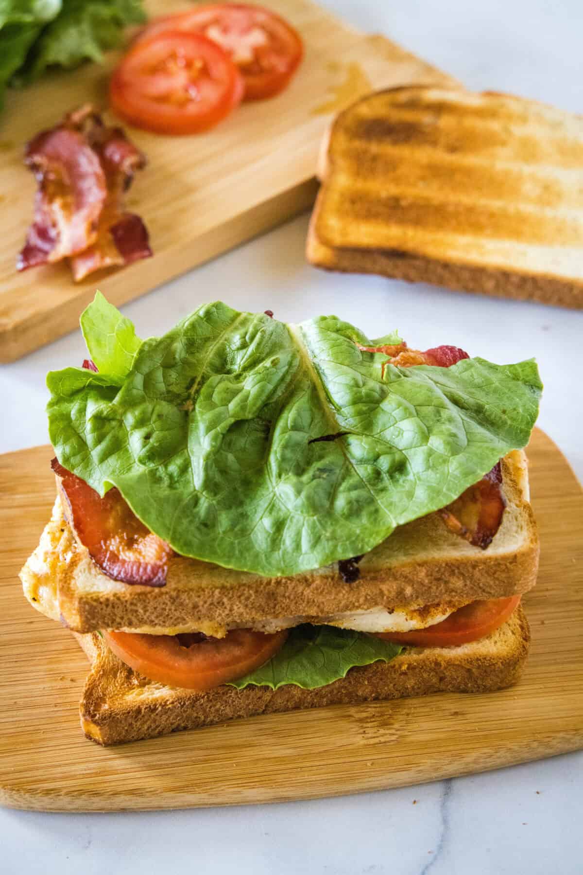 The second bread slice, bacon, and lettuce added to a chicken club sandwich on a wooden cutting board.