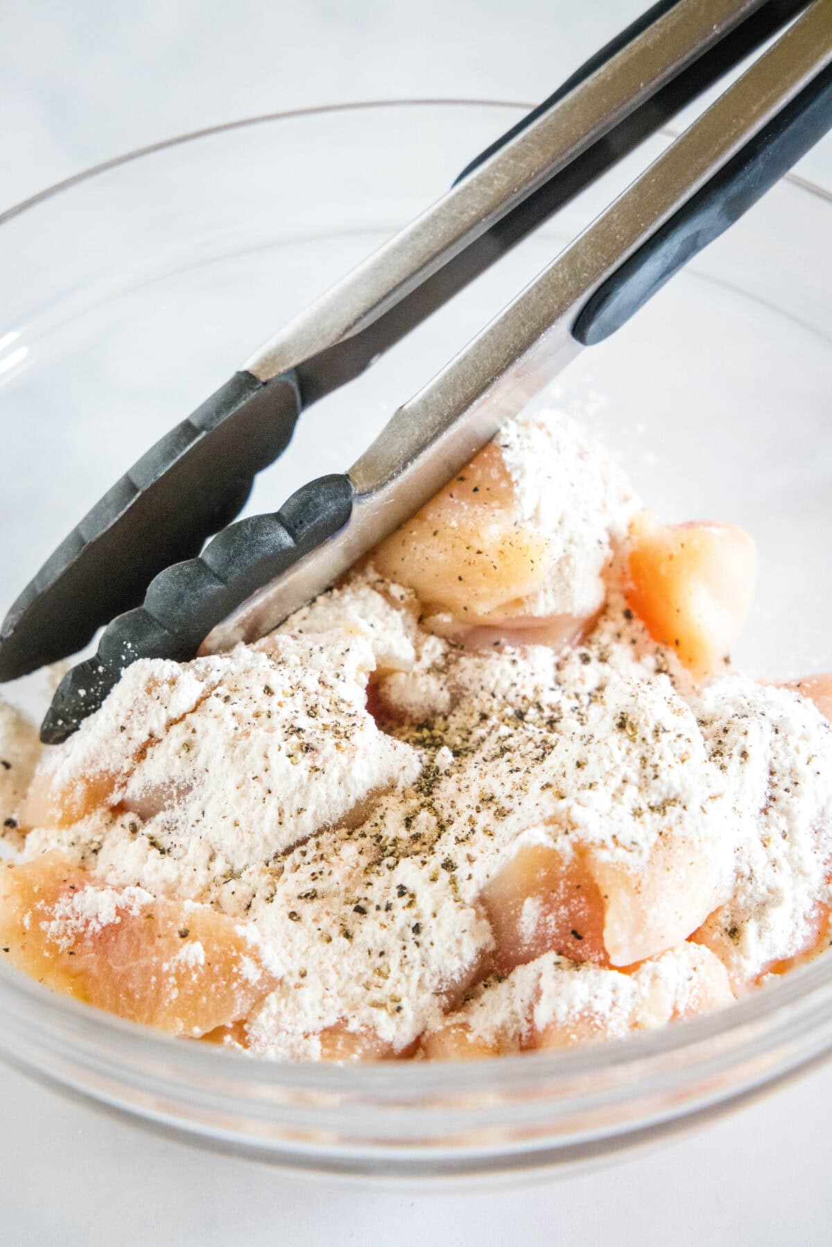 This chicken scampi recipe is a quick, easy chicken pasta with juicy chicken bites cooked in a buttery white wine sauce with garlic and lemon. Flour added to chicken pieces in a glass bowl, with tongs.