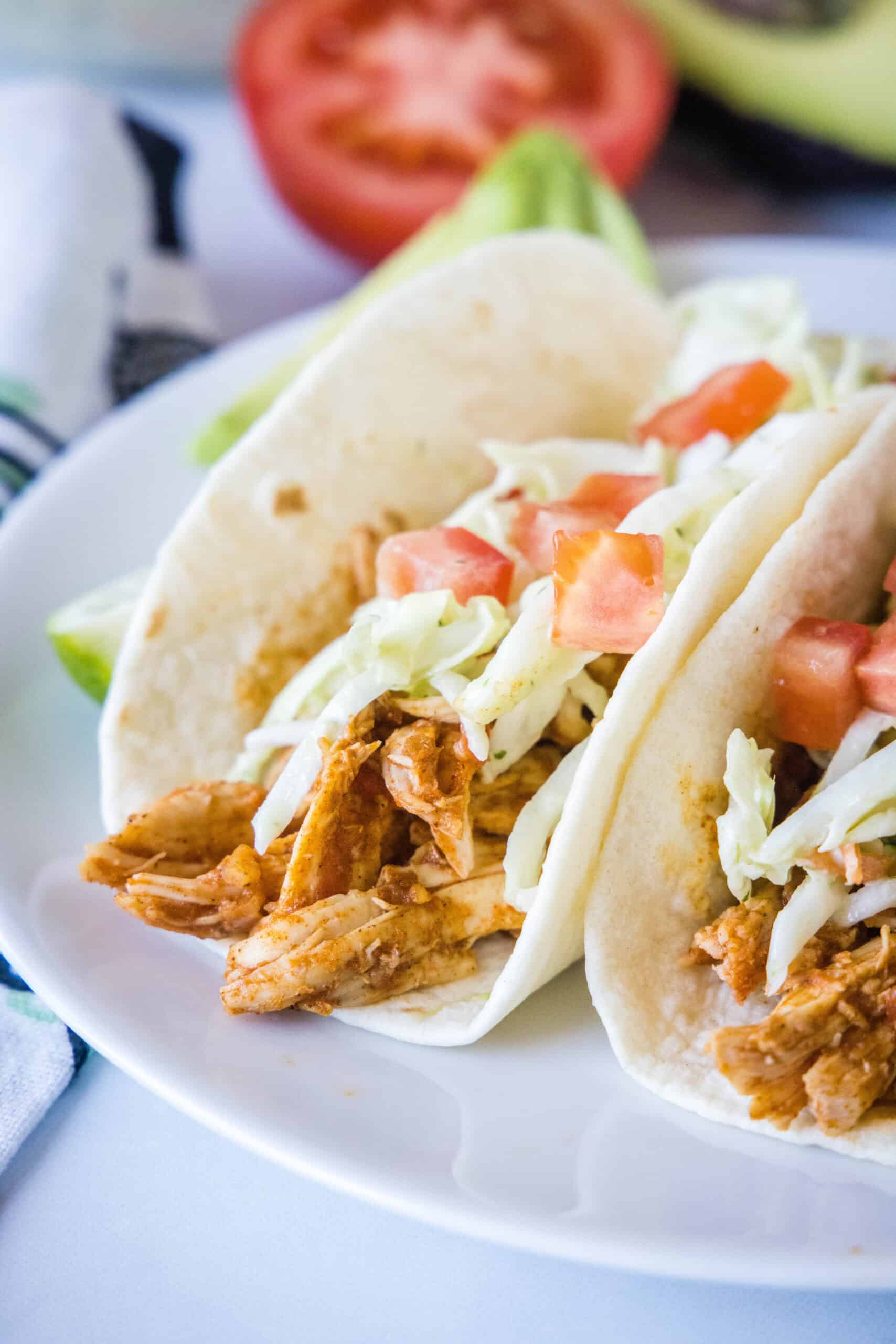 Juicy rotisserie chicken tacos are the best shortcut on taco night! Make delicious, easy chicken tacos in 15 minutes with only 3 ingredients. Two rotisserie chicken tacos topped with Mexican coleslaw and diced tomatoes on a white plate.