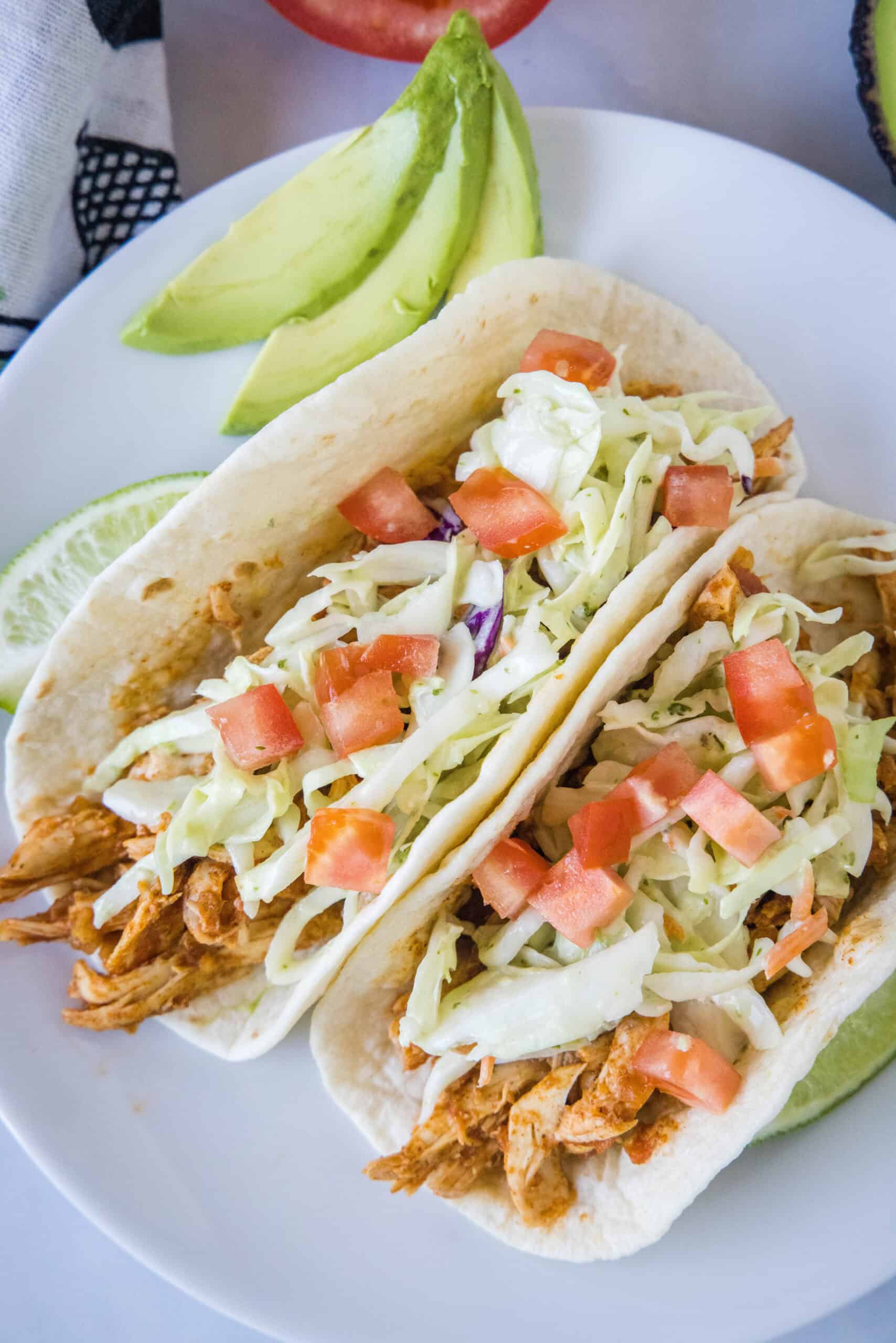 Juicy rotisserie chicken tacos are the best shortcut on taco night! Make delicious, easy chicken tacos in 15 minutes with only 3 ingredients. Overhead view of two rotisserie chicken tacos topped with Mexican coleslaw and diced tomatoes on a white plate, next to sliced avocado.