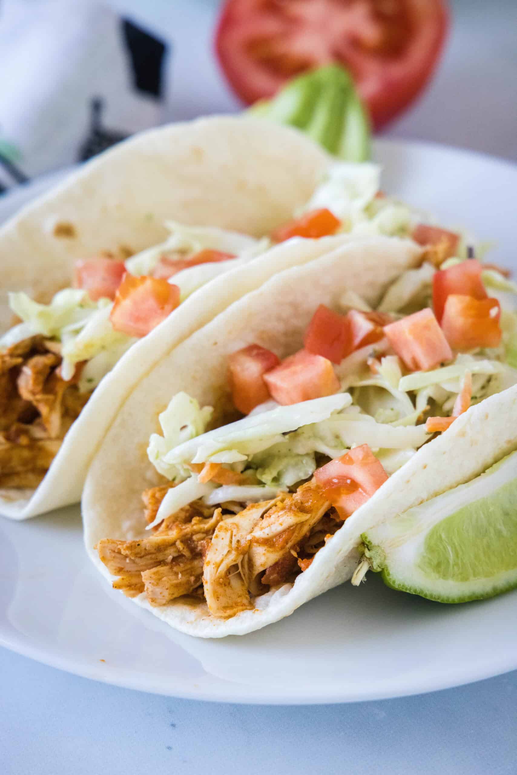 Juicy rotisserie chicken tacos are the best shortcut on taco night! Make delicious, easy chicken tacos in 15 minutes with only 3 ingredients. Two rotisserie chicken tacos topped with Mexican coleslaw and diced tomatoes on a white plate, next to a lime wedge.