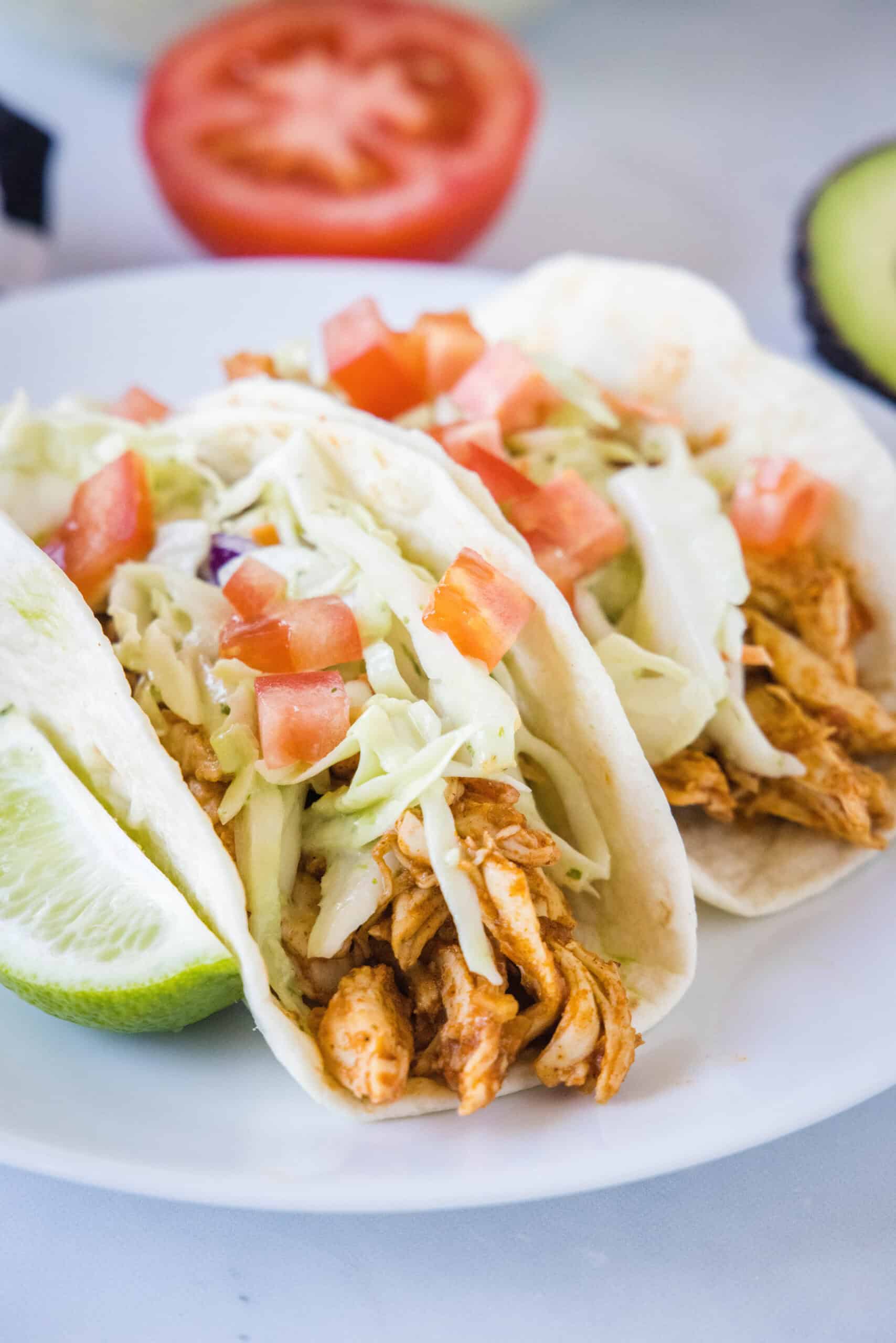 Juicy rotisserie chicken tacos are the best shortcut on taco night! Make delicious, easy chicken tacos in 15 minutes with only 3 ingredients. Two rotisserie chicken tacos topped with Mexican coleslaw and diced tomatoes on a white plate, next to a lime wedge.