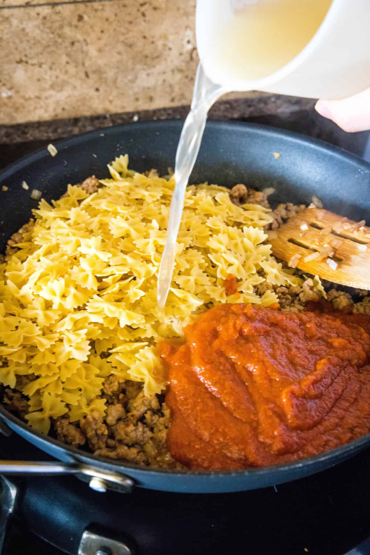 pouring chicken broth into a skillet with dried pasta and tomato sauce